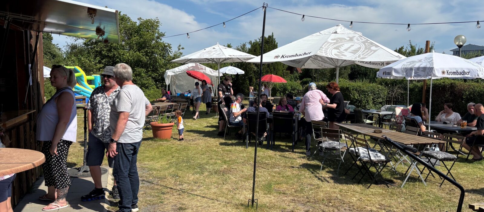 Gäste an Biertischen auf der Festwiese vor dem Vereinsheim, dazwischen Sonnenschirme und eine Wimpelkette bei blauem Himmel